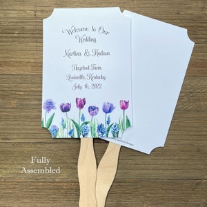 Op de afbeelding: Een witte bruiloftswaaier met een bloemenontwerp met paarse en blauwe tulpen. De waaier is volledig gemonteerd en heeft een houten handvat. De tekst op de waaier luidt "Welcome to Our Wedding Martina & Hudson Hazelnut Farm Louisville, Kentucky July 16, 2022".