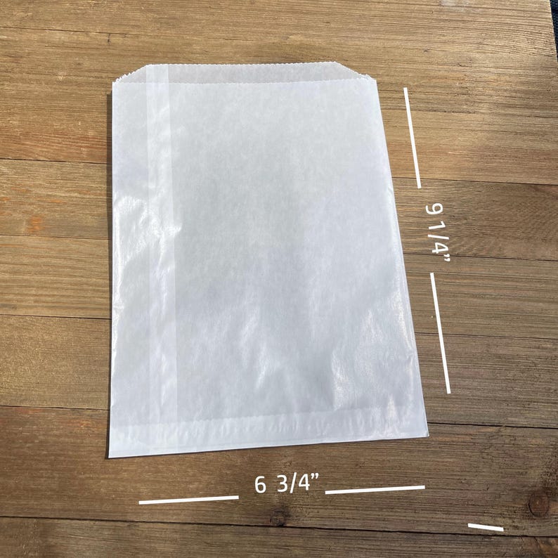 Puede incluir: Una bolsa de papel blanca, que mide 23,5 cm de alto y 17,1 cm de ancho, est&aacute; sobre una superficie de madera. La bolsa es rectangular con una parte superior doblada y una costura lateral. El papel parece ligeramente transl&uacute;cido.