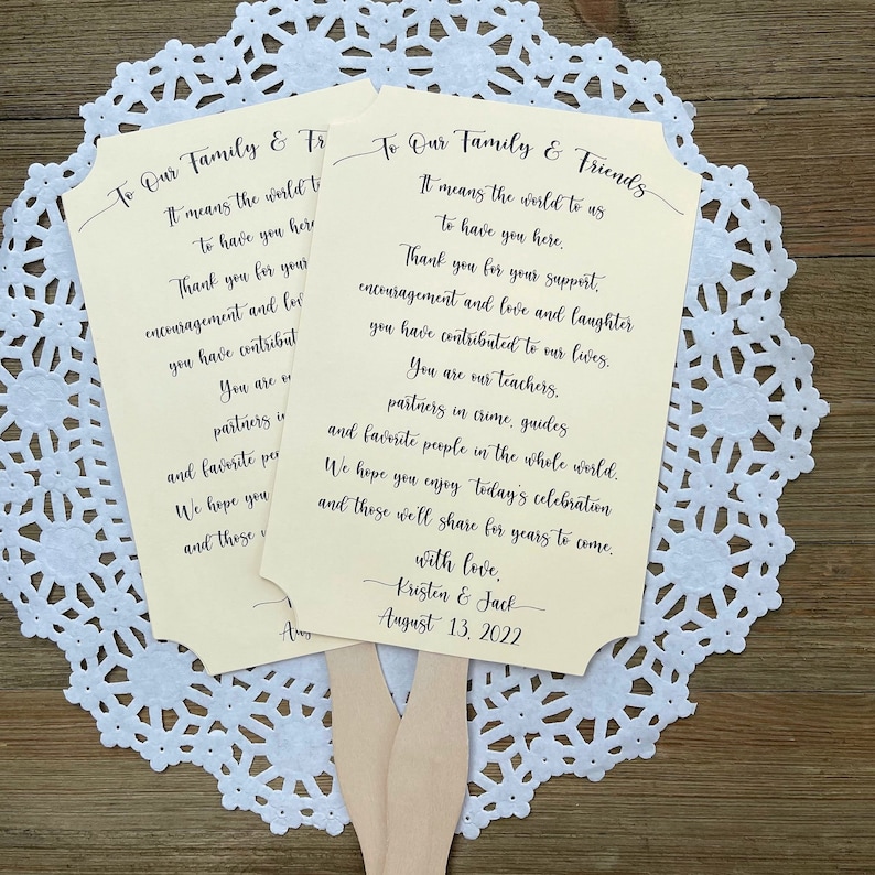 May include: Two ivory-colored wedding fans with black text. The text reads "To Our Family & Friends" and includes a message of thanks to guests for their support and love. The fans are decorated with a simple black border and are held together with a wooden stick.