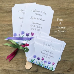 Op de afbeelding: Huwelijksvaandels met een bloemenontwerp met paarse tulpen en blauwe bloemen. De vaandels zijn wit met een houten handvat en een eraan bevestigd lint. De tekst op het vaandel luidt "Lucky In Love... Martina & Hudson July 16, 2022".
