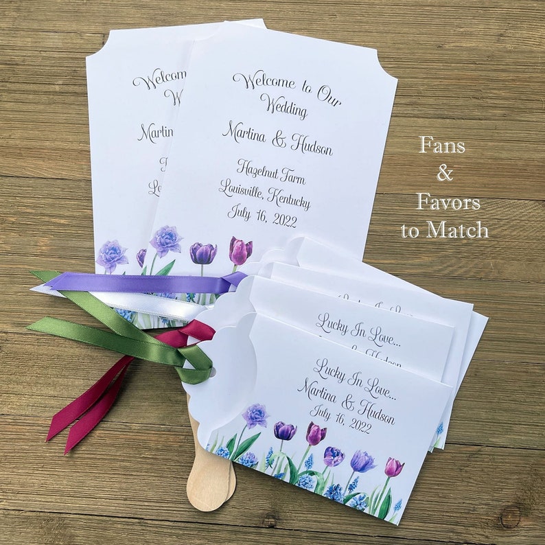 Op de afbeelding: Huwelijksvaandels met een bloemenontwerp met paarse tulpen en blauwe bloemen. De vaandels zijn wit met een houten handvat en een eraan bevestigd lint. De tekst op het vaandel luidt "Lucky In Love... Martina & Hudson July 16, 2022".