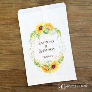 Pode incluir: Saco de papel branco com um design floral com girass&oacute;is e vegeta&ccedil;&atilde;o. O saco tem os nomes "Raymond & Shannon" e a data "08-04-XX" impressos. O saco &eacute; da Abbey and Izzie Designs.
