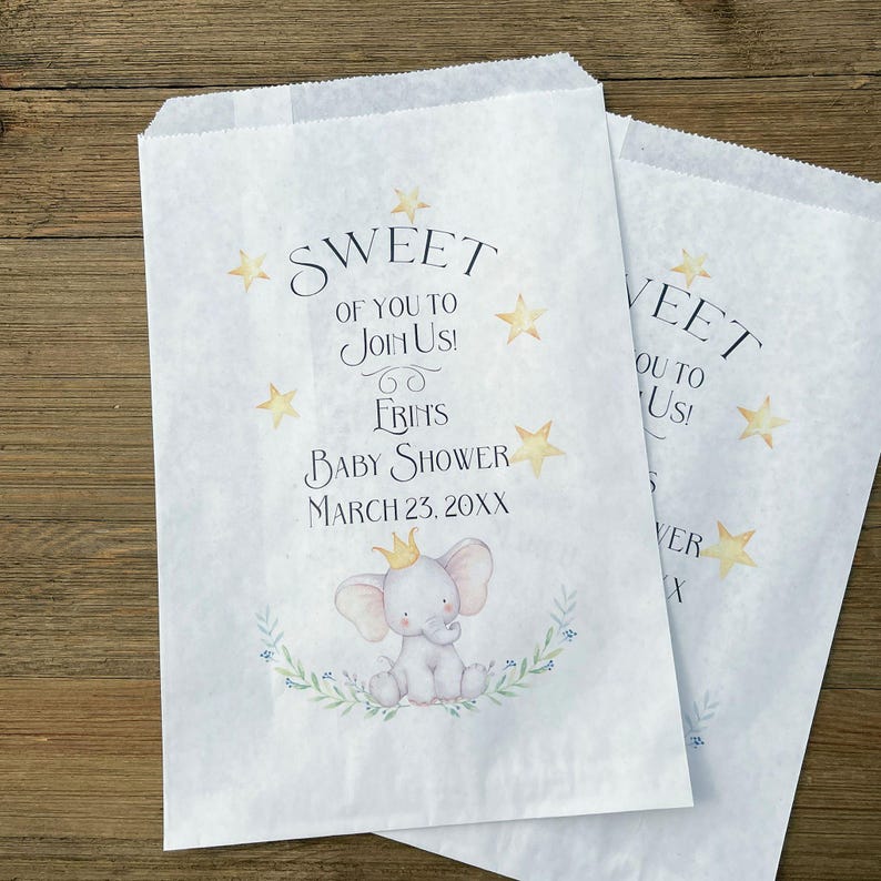 May include: Two white paper treat bags with the text "SWEET OF YOU TO JOIN US!" and "FRIN'S BABY SHOWER MARCH 23, 20XX". The bags feature a watercolor illustration of a baby elephant wearing a crown, surrounded by a floral wreath and gold stars.