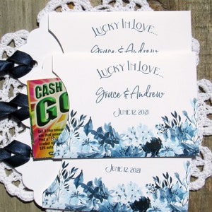 May include: Two white wedding favor cards with blue floral designs and the text "Lucky in Love... Grace & Andrew June 12, 2021". The top card has a small, partially visible, green and white "Cash Go" scratch-off ticket.