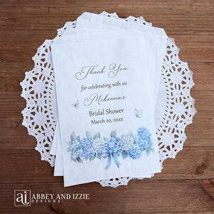 K&ouml;nnte beinhalten: Wei&szlig;e Papiert&uuml;ten mit blauem Hortensienblumendesign und dem Text "Thank You for celebrating with us McKenna's Bridal Shower March 20, 20xx". Die T&uuml;ten liegen auf einer wei&szlig;en Spitzendecke.