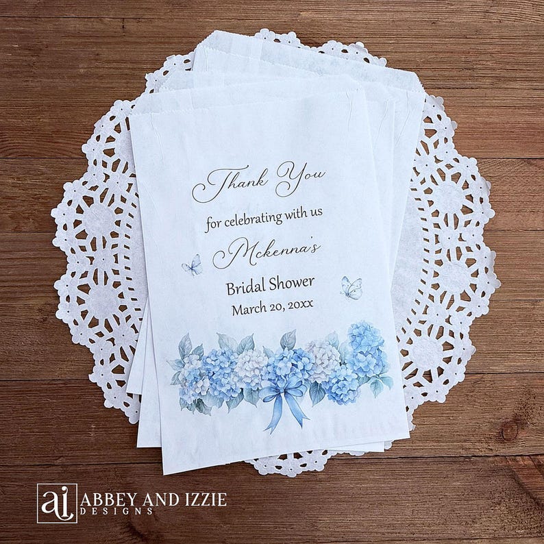 K&ouml;nnte beinhalten: Wei&szlig;e Papiert&uuml;ten mit blauem Hortensienblumendesign und dem Text "Thank You for celebrating with us McKenna's Bridal Shower March 20, 20xx". Die T&uuml;ten liegen auf einer wei&szlig;en Spitzendecke.