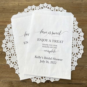 May include: White paper favor bags with black text that reads "love is sweet, Enjoy a Treat, Thank you for making my day complete, Kelly's Bridal Shower, July 16, 2022".