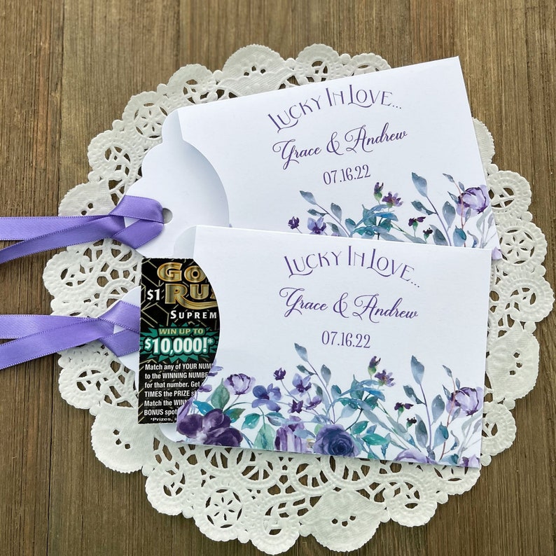May include: Two white envelopes with a floral design in shades of purple and blue. The envelopes are decorated with the text "Lucky in Love... Grace & Andrew 07.16.22". One envelope has a scratch-off lottery ticket peeking out from the top.