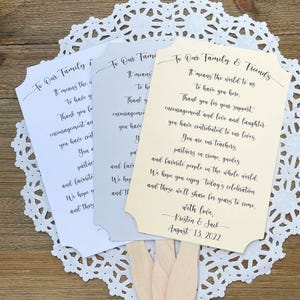 May include: Three wedding fan programs with a light brown wooden handle. The programs are printed with a message thanking guests for their support and contribution to the couple's lives. The programs are in white, light gray, and light yellow. The yellow program has the text "To Our Family & Friends" and the date "August 13, 2022".
