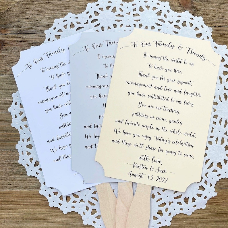May include: Three wedding fan programs with a light brown wooden handle. The programs are printed with a message thanking guests for their support and contribution to the couple's lives. The programs are in white, light gray, and light yellow. The yellow program has the text "To Our Family & Friends" and the date "August 13, 2022".