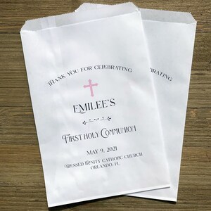 May include: Two white paper treat bags with text. The bags read "Thank you for celebrating" above a pink cross and the name "Emilee's". Below is "First Holy Communion" and the date "May 9, 2021".