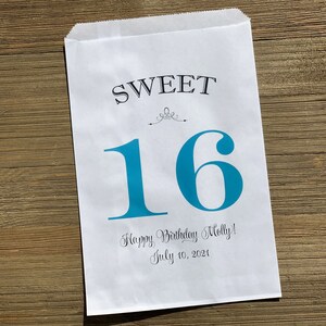 Op de afbeelding: Witte papieren cadeauzak met de tekst "SWEET 16" in zwart en blauwgroen. De zak bevat ook de tekst "Happy Birthday Molly! July 10, 2021". De zak ligt op een houten ondergrond.