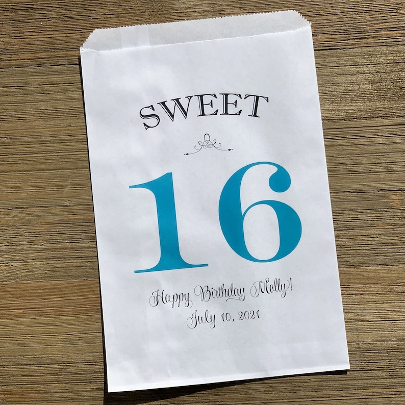 Op de afbeelding: Witte papieren cadeauzak met de tekst "SWEET 16" in zwart en blauwgroen. De zak bevat ook de tekst "Happy Birthday Molly! July 10, 2021". De zak ligt op een houten ondergrond.