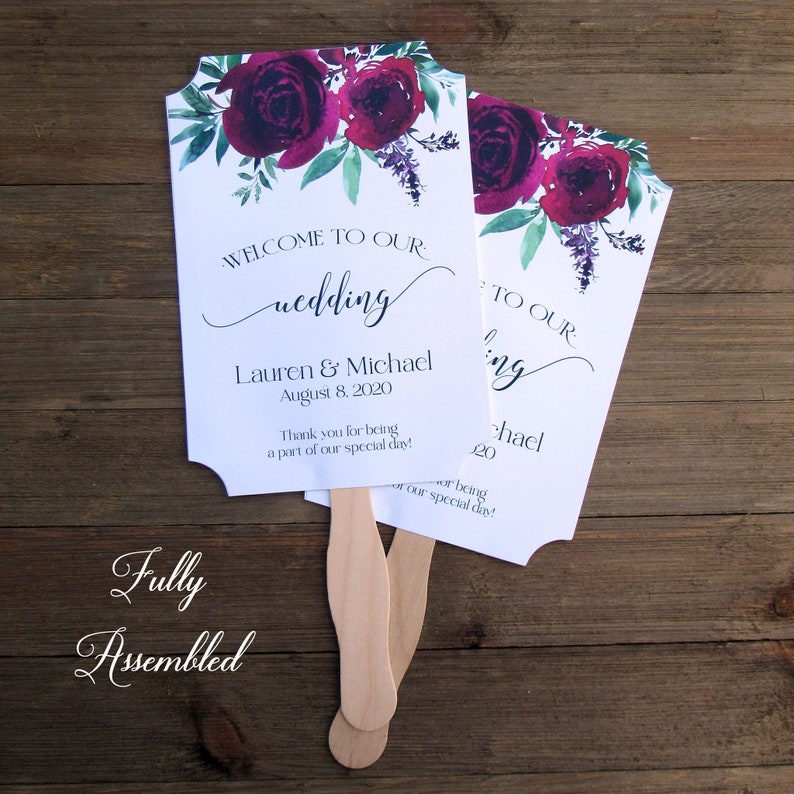 May include: Two white wedding fans with burgundy floral designs and green leaves. The fans have wooden handles and the text "Welcome to our wedding" and "Lauren & Michael August 8, 2020" printed on them.