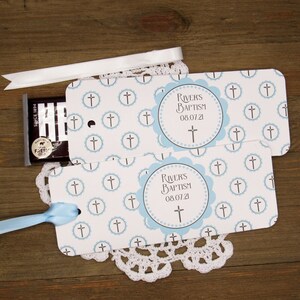 K&ouml;nnte beinhalten: Zwei blaue und wei&szlig;e Geschenkanh&auml;nger mit einem Kreuzmuster und einem blauen Kreis mit dem Text "RIVER'S BAPTISM 08.07.21" und einem Kreuz darin. Die Anh&auml;nger sind mit blauem und wei&szlig;em Band gebunden.