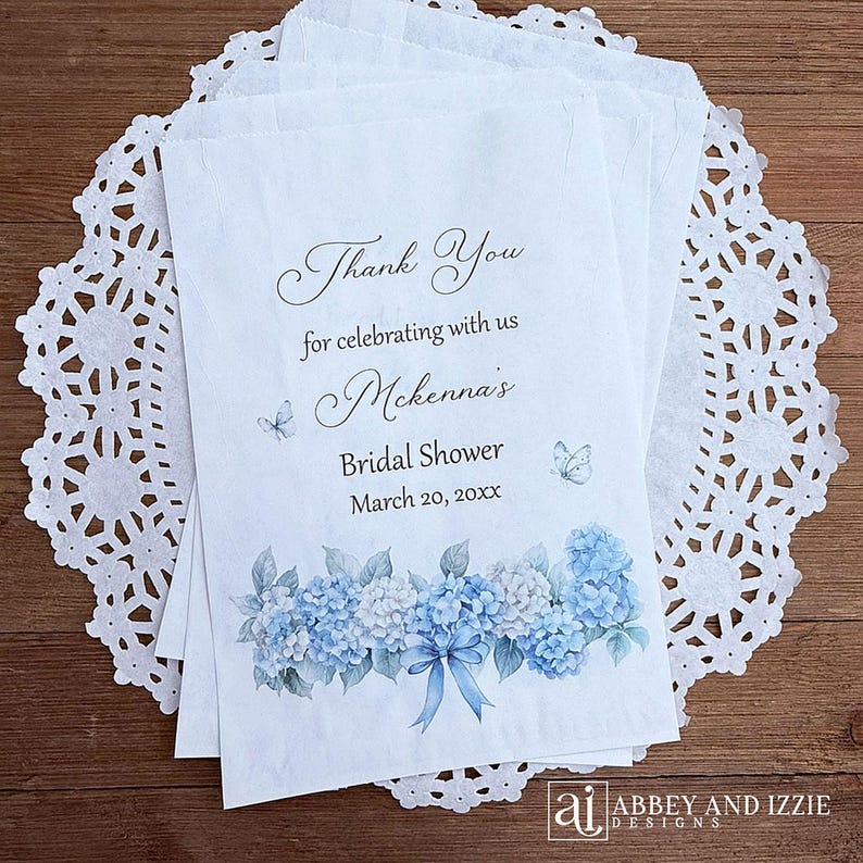 K&ouml;nnte beinhalten: Wei&szlig;e Papiert&uuml;ten mit dem Text "Thank You" und "Mckenna's Bridal Shower March 20, 20xx". Die T&uuml;ten sind mit blau-wei&szlig;en Hortensienbl&uuml;ten und Schmetterlingen verziert. Die T&uuml;ten liegen auf einer wei&szlig;en Spitzendecke.