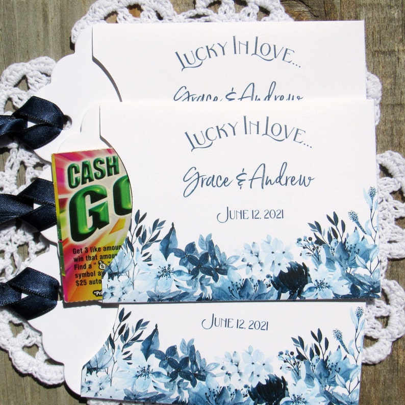 May include: Two white wedding favor cards with blue floral designs and the text "Lucky in Love... Grace & Andrew June 12, 2021". The top card has a small, partially visible, green and white "Cash Go" scratch-off ticket.