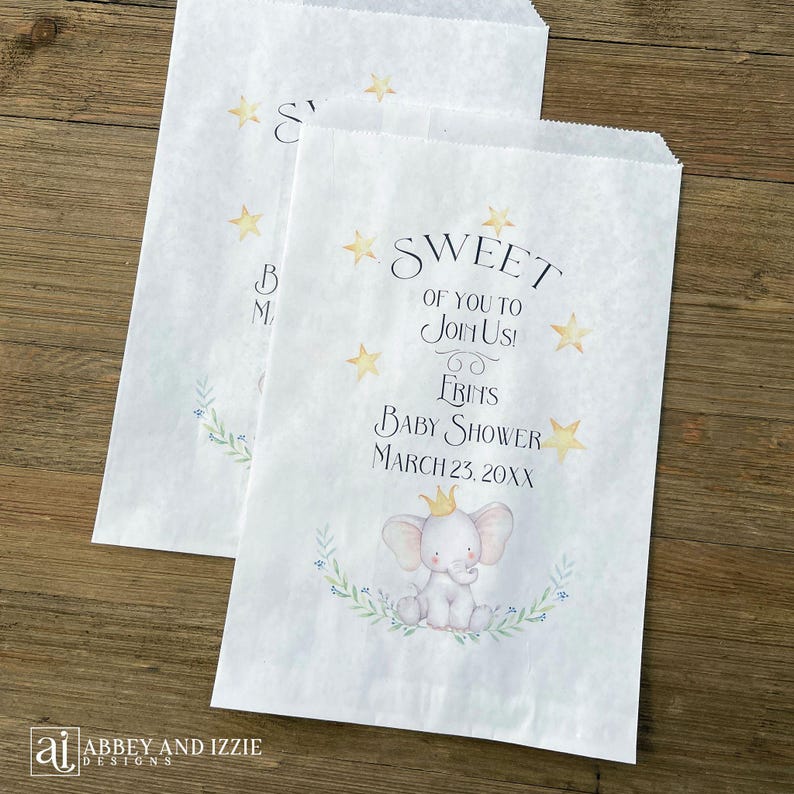 May include: Two white paper favor bags with a baby shower theme. The bags feature text that reads "Sweet of you to join us! Frins Baby Shower March 23, 20XX" and a watercolor illustration of a baby elephant wearing a crown.