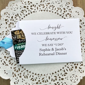 K&ouml;nnte beinhalten: Eine wei&szlig;e Karte mit schwarzem Text, der lautet: "Heute Abend feiern wir mit euch. Morgen sagen wir "Ja" Sophie & Jacob's Rehearsal Dinner". Ein blaues Band ist an ein kleines Rubbellos befestigt, das durch einen Ausschnitt in der Karte teilweise sichtbar ist.