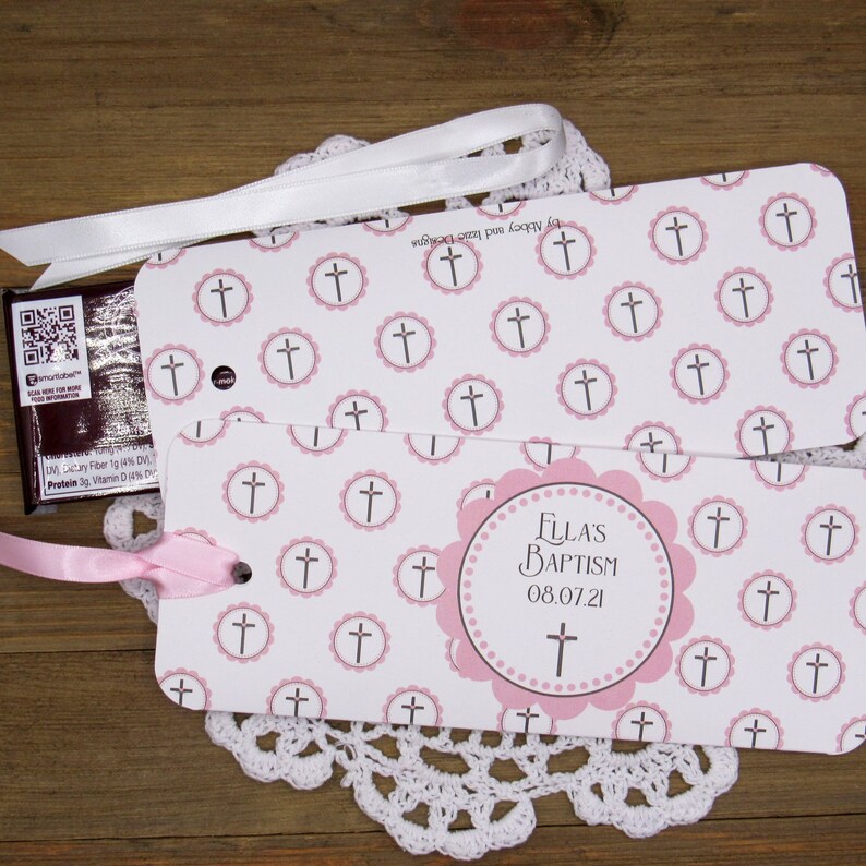 May include: A white and pink favor box with a pink ribbon. The box has a cross pattern and the text "FLLA'S BAPTISM 08.07.21".