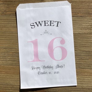 Op de afbeelding: Witte papieren cadeauzak met de tekst "SWEET 16" in zwarte en roze letters. De zak bevat ook de tekst "Happy Birthday Alexis! October 10, 2020". De zak ligt op een houten oppervlak.