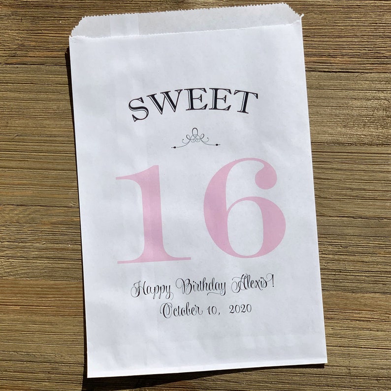 Op de afbeelding: Witte papieren cadeauzak met de tekst "SWEET 16" in zwarte en roze letters. De zak bevat ook de tekst "Happy Birthday Alexis! October 10, 2020". De zak ligt op een houten oppervlak.