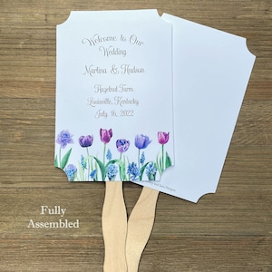 Op de afbeelding: Een witte bruiloftswaaier met een bloemenontwerp met paarse en blauwe tulpen. De waaier is volledig gemonteerd en heeft een houten handvat. De tekst op de waaier luidt "Welcome to Our Wedding Martina & Hudson Hazelnut Farm Louisville, Kentucky July 16, 2022".