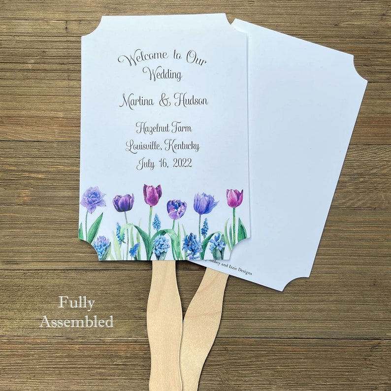Op de afbeelding: Een witte bruiloftswaaier met een bloemenontwerp met paarse en blauwe tulpen. De waaier is volledig gemonteerd en heeft een houten handvat. De tekst op de waaier luidt "Welcome to Our Wedding Martina & Hudson Hazelnut Farm Louisville, Kentucky July 16, 2022".