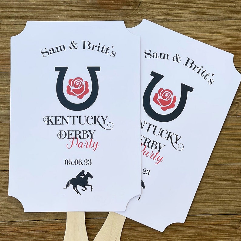 May include: Two white paper fans with black and red horseshoe and rose designs. The text on the fans reads "Sam & Britt's Kentucky Derby Party 05.06.23".