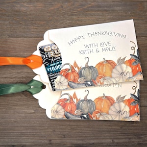 May include: Two white cardstock lottery ticket holders with scalloped edges and orange and green satin ribbons. The cards feature a watercolor pumpkin illustration and the text "HAPPY THANKSGIVING WITH LOVE, KEITH & MOLLY" printed in black.