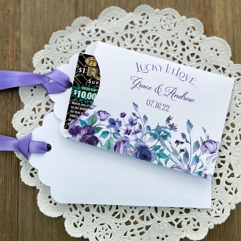 May include: White favor bag with a purple floral design and the text "LUCKY IN LOVE... Grace & Andrew 07.16.22". A lottery ticket is partially visible inside the bag.