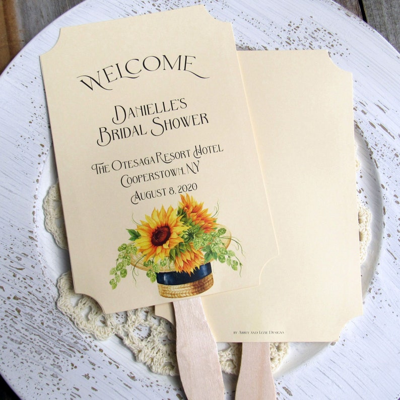 May include: A cream-colored bridal shower fan with a sunflower design and the text "WELCOME DANIELLE'S BRIDAL SHOWER THE OTESAGA RESORT HOTEL COOPERSTOWN, NY AUGUST 8, 2020".