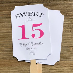 Op de afbeelding: Een witte en roze papieren waaier met de tekst "SWEET 15 Bridget's Quincea&ntilde;era July 10, 2021" en een decoratief ontwerp. De waaier is bevestigd aan een houten stok.