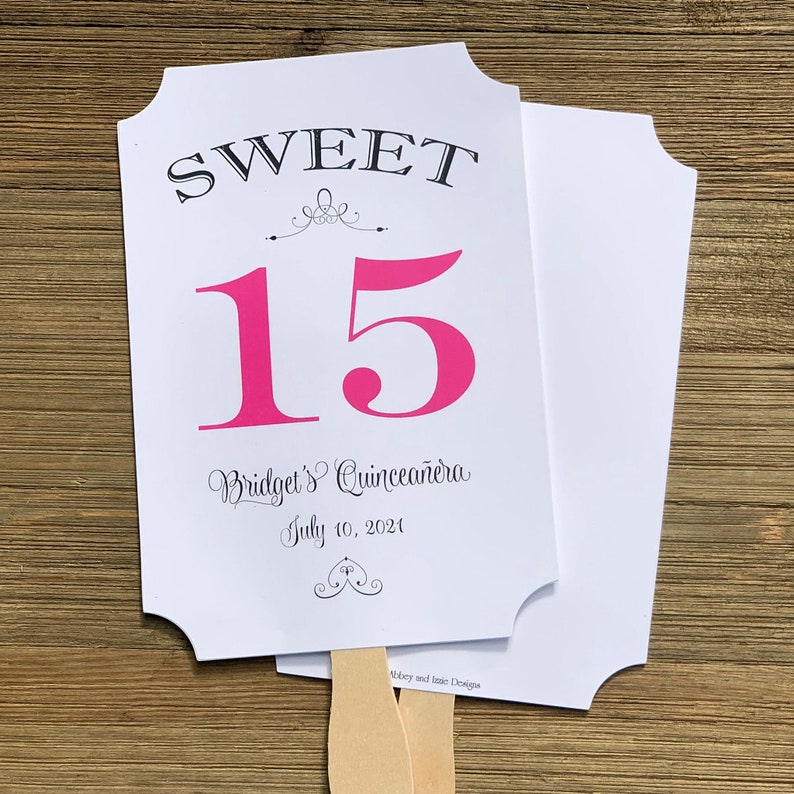 Op de afbeelding: Een witte en roze papieren waaier met de tekst "SWEET 15 Bridget's Quincea&ntilde;era July 10, 2021" en een decoratief ontwerp. De waaier is bevestigd aan een houten stok.