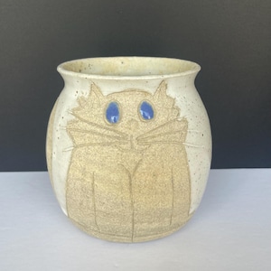 Puede incluir: Un jarrón de cerámica blanca con un esmalte moteado presenta un diseño en relieve de un gato con ojos azules. El jarrón es redondo y tiene una abertura ancha.