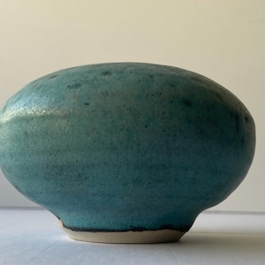 May include: A handcrafted, teal-colored ceramic bowl with a textured surface. The bowl has a rounded shape with a slightly flared base and a contrasting off-white rim. The bowl is suitable for serving or display.