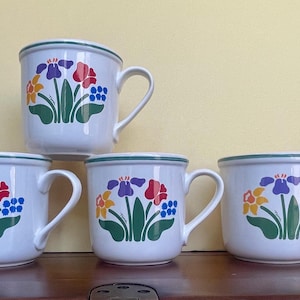 May include: Four white ceramic mugs with green rims and floral designs. Each mug features a different arrangement of red, yellow, blue, and green flowers.