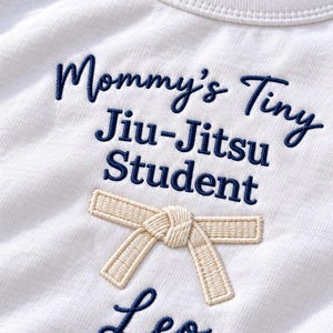 Op de afbeelding: Een wit katoenen shirt met donkerblauw geborduurde tekst die Mommy's Tiny Jiu-Jitsu Student en de naam Leo leest. Onder de tekst bevindt zich een geborduurde beige vechtsportgordel in een knoop, gecentreerd op de borst van het kledingstuk.