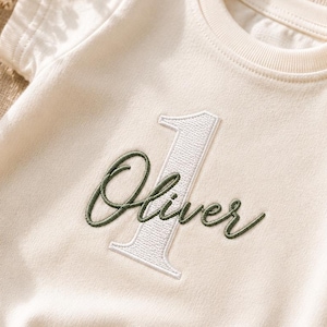 Può includere: Un body per neonati a maniche corte color crema con il numero uno ricamato in bianco e il nome Oliver cucito in corsivo verde scuro al centro. Il capo presenta un girocollo a coste e chiusure a pressione nella parte inferiore.