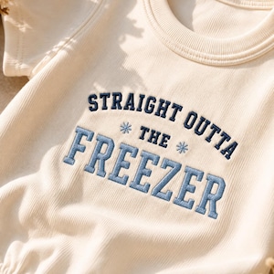 Może przedstawiać: Kremowe body z krótkim rękawem wykonane z prążkowanej dzianiny, ozdobione ciemnoniebieskim haftem o treści STRAIGHT OUTTA THE FREEZER, z dwoma małymi niebieskimi ikonami płatków śniegu po bokach słowa THE. Ubranie posiada okrągły dekolt i metalowe zatrzaski.