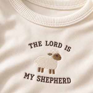 Könnte beinhalten: Ein cremeweißes Rippshirt mit einer Stickerei eines kleinen Schafs in Creme und Braun. Der Text "THE LORD IS MY SHEPHERD" ist in braunen Großbuchstaben oberhalb und unterhalb der Schaf-Illustration auf der Brust eingestickt.