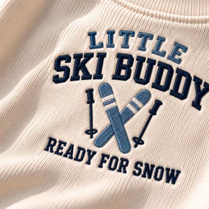 May include: A cream-colored ribbed sweatshirt featuring dark blue embroidered text that reads LITTLE SKI BUDDY and READY FOR SNOW, with a central graphic of two light blue skis and two dark blue ski poles stitched onto the fabric.
