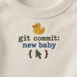 Könnte beinhalten: Ein cremefarbenes, geripptes Rundhals-Shirt mit einer Stickerei eines gelben Quietscheentchens über dem Text "git commit: new baby" in Dunkelgrün und Blau, gefolgt von einem grünen, verpixelten Mauszeiger zwischen zwei braunen geschweiften Klammern.