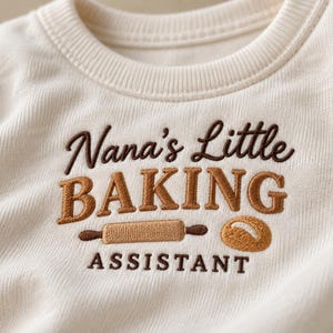 Könnte beinhalten: Ein cremefarbenes Sweatshirt mit Rundhalsausschnitt und dunkelbrauner sowie hellbrauner Stickerei. Der Text lautet Nana's Little BAKING ASSISTANT, ergänzt durch ein gesticktes Nudelholz und ein kleines Brot im Brustbereich des Kleidungsstücks.