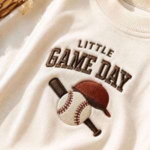 Op de afbeelding: Een crèmekleurig sweatshirt met ronde hals en geborduurde tekst LITTLE GAME DAY in donkerbruine blokletters. Onder de tekst staat een geborduurde afbeelding van een donkerbruine honkbalknuppel, een witte honkbal en een roestkleurige pet.