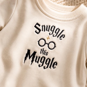 Può includere: Una felpa girocollo color crema con ricamo nero che recita Snuggle this Muggle. Il design include un paio di occhiali circolari neri e una piccola stella dorata sopra gli occhiali, con un dettaglio a forma di fulmine sulla lettera M.