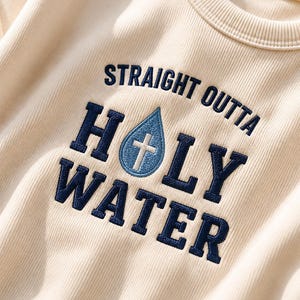 May include: A cream-colored ribbed sweatshirt featuring dark blue embroidered text that reads STRAIGHT OUTTA HOLY WATER. The letter O in HOLY is replaced by a light blue embroidered water droplet containing a white cross symbol.
