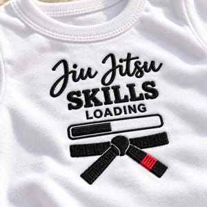 May include: A white short-sleeved shirt featuring black embroidered text that reads Jiu Jitsu SKILLS LOADING. Below the text is a loading bar graphic and a black martial arts belt with a red stripe detail, all rendered in embroidery.