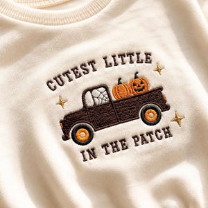 Peut inclure: Un sweat-shirt crème avec une broderie marron foncé représentant un pick-up transportant deux citrouilles. Le texte indique CUTEST LITTLE IN THE PATCH, accompagné de petites étoiles dorées brodées. Le camion a des roues orange et une toile d'araignée.