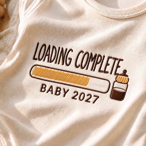 May include: A cream-coloured ribbed baby bodysuit featuring brown embroidered text that reads LOADING COMPLETE and BABY 2027. The design includes a yellow progress bar and a brown baby bottle icon with a yellow nipple, centred on the chest.